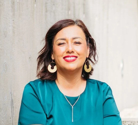 Woman smiling, wearing a teal top and gold earrings, against a gray wall.
