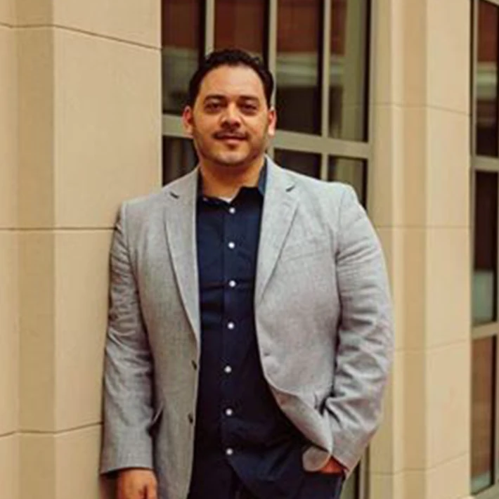 Man in gray blazer and navy shirt leaning against a building.