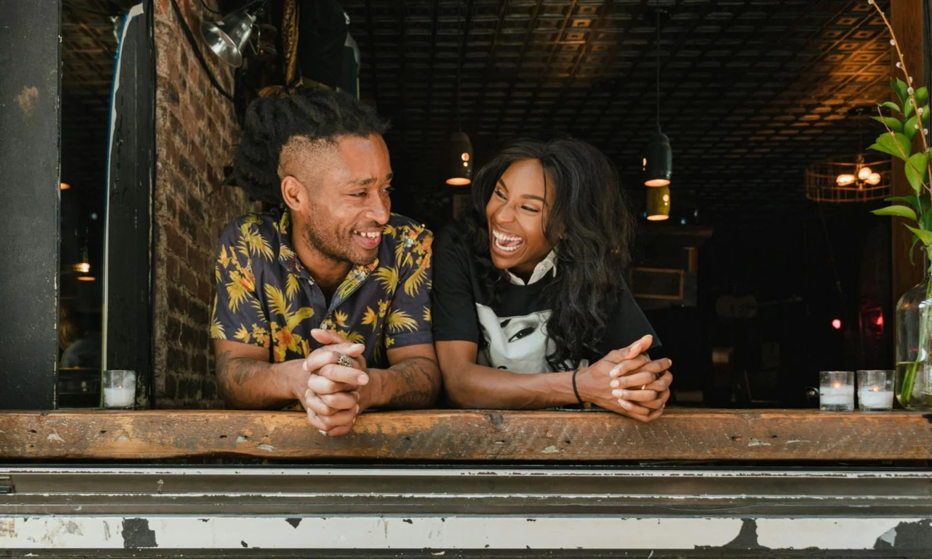 Two people laughing, leaning on a wooden ledge. One wears a patterned shirt, the other a graphic t-shirt. Dark interior setting.