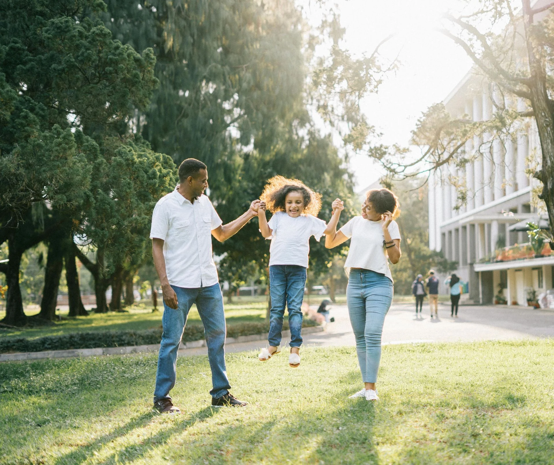Family holding hands, child jumping in sunny park, green grass, trees, and a building in the background.
