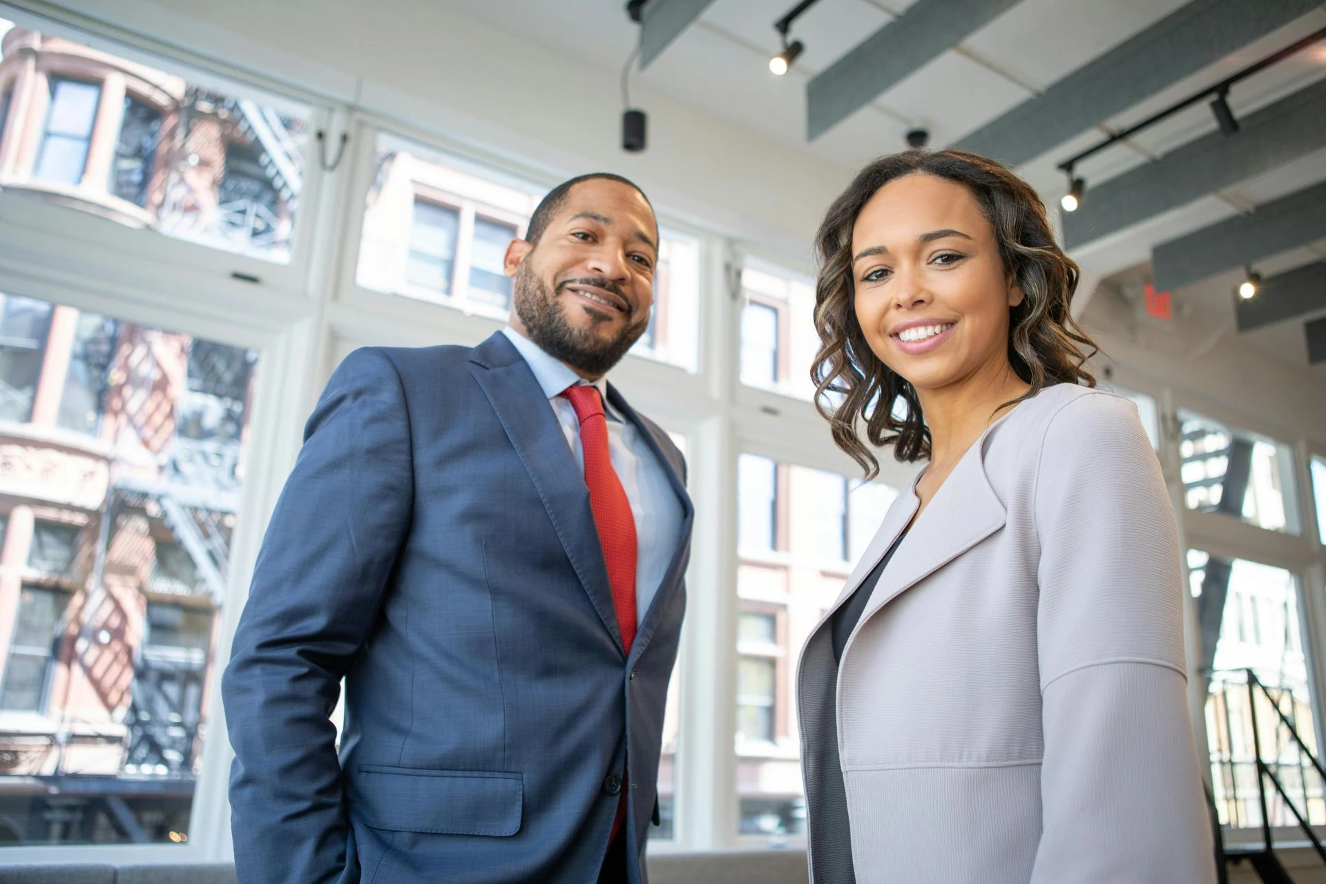 Man in suit with a red tie and woman in a gray blazer smiling in an office with large windows.