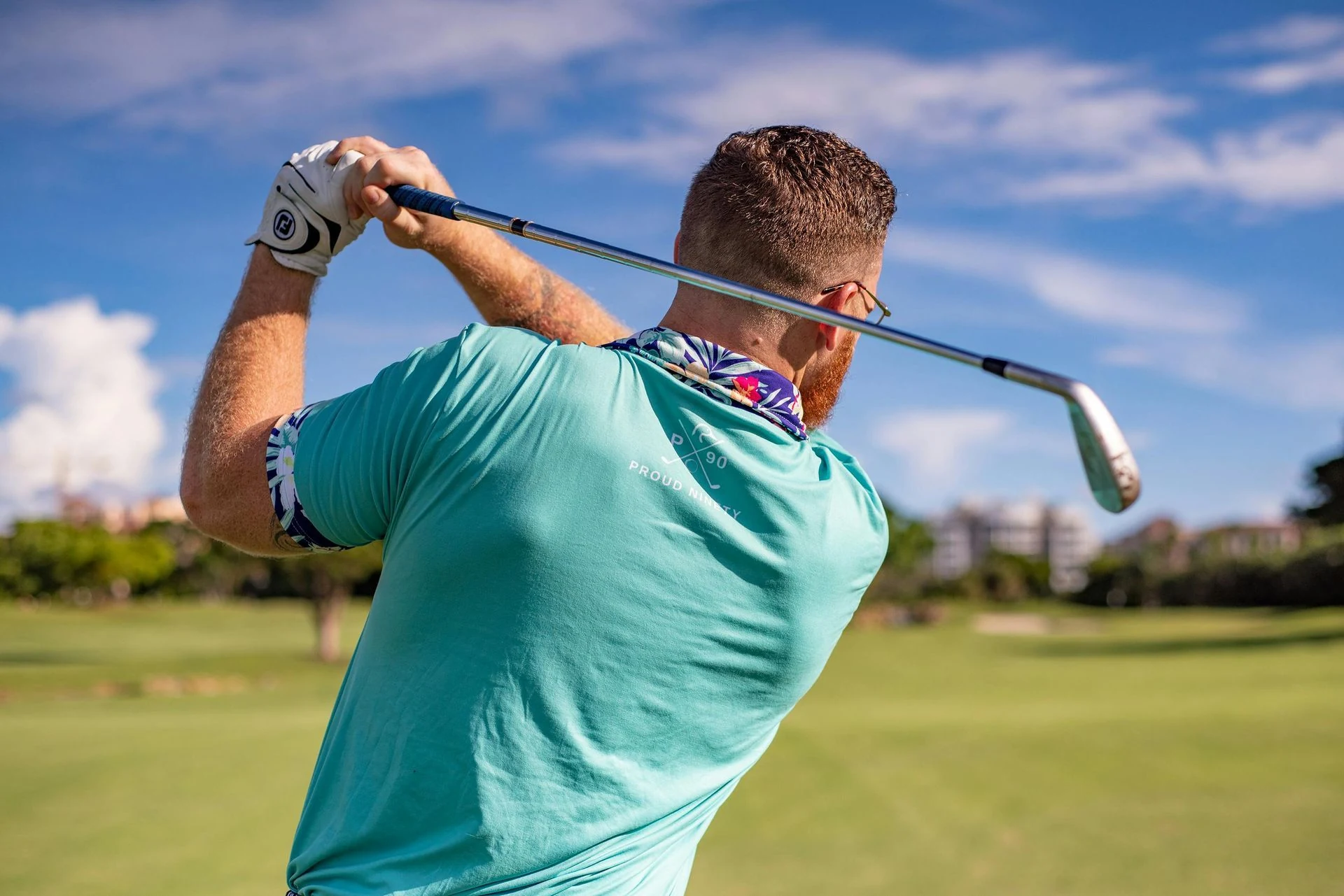 Golfer swinging a club on a green course, blue sky, teal shirt, white glove.