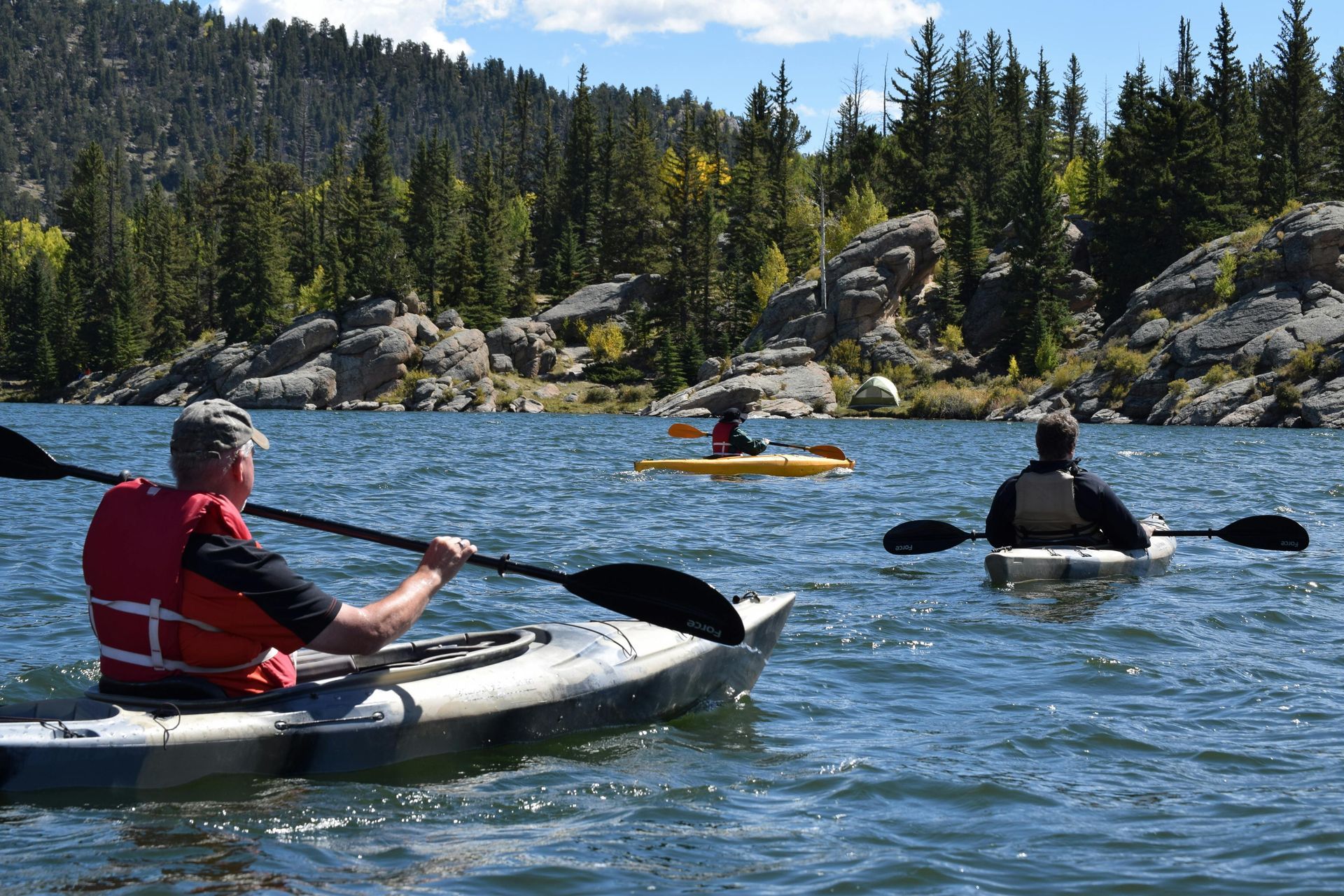 Three people kayaking on a lake surrounded by trees and rocks on a sunny day.