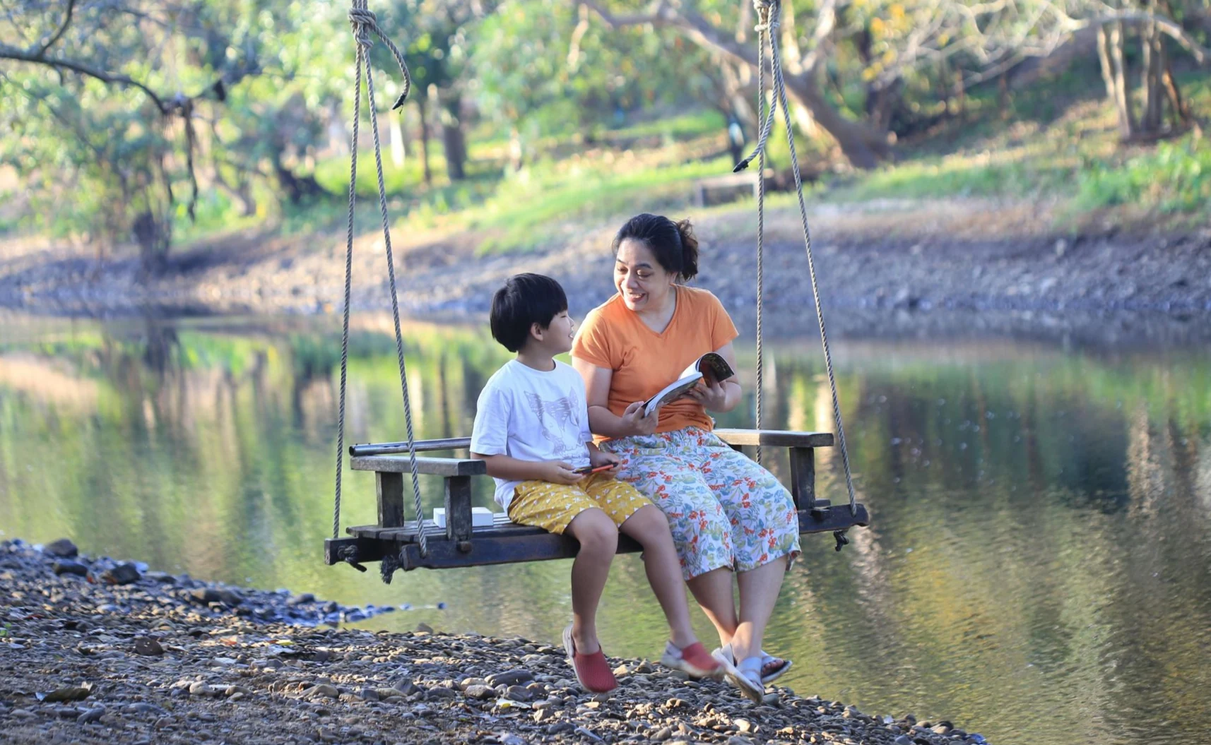 Woman and child sit on swing by a calm river, conversing. Trees line the background.