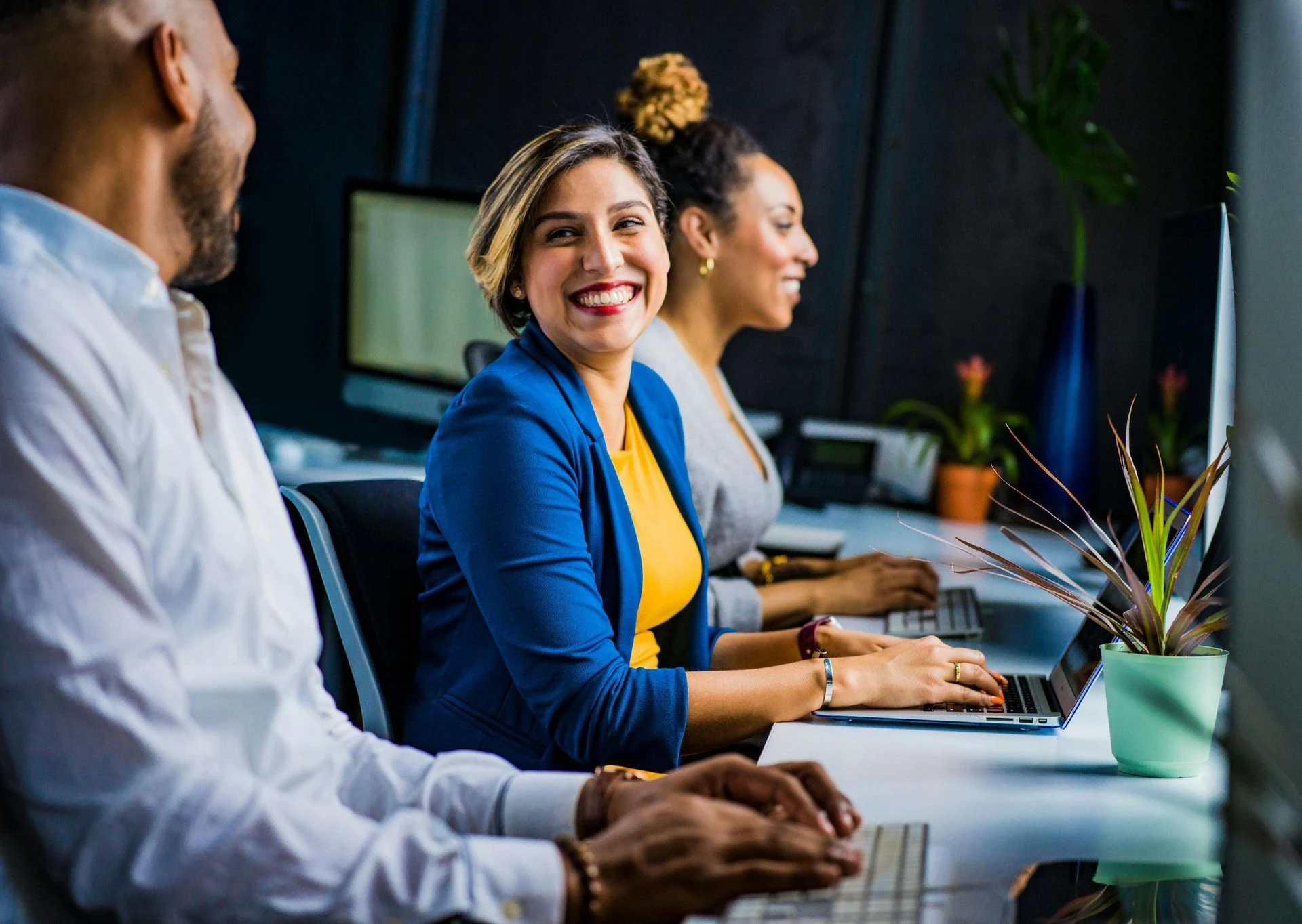 Three people working at desks; a woman smiles at the camera, wearing a blue jacket, yellow shirt; a computer and plants are visible.