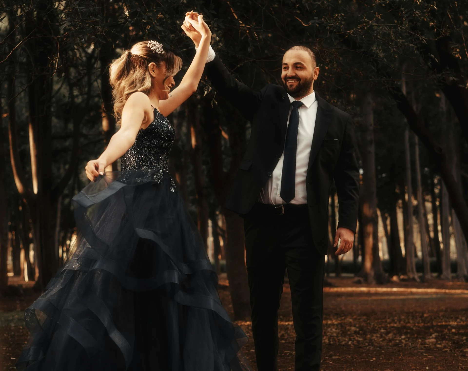 Couple dancing outdoors; woman in blue gown, man in suit, holding hands, trees in background.
