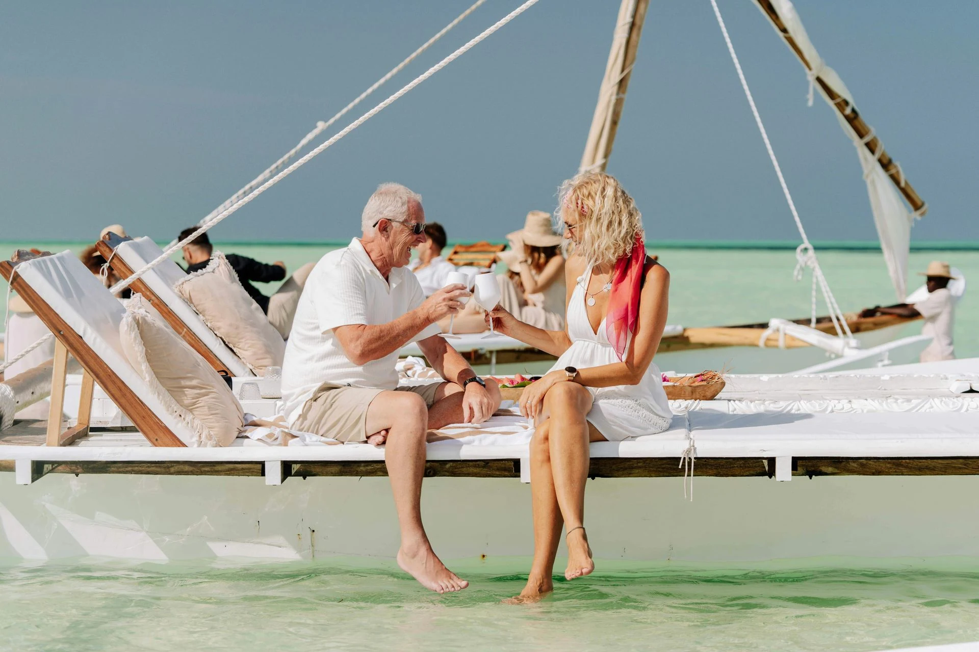 Man and woman toasting glasses on a white platform over turquoise water. Others lounge nearby.