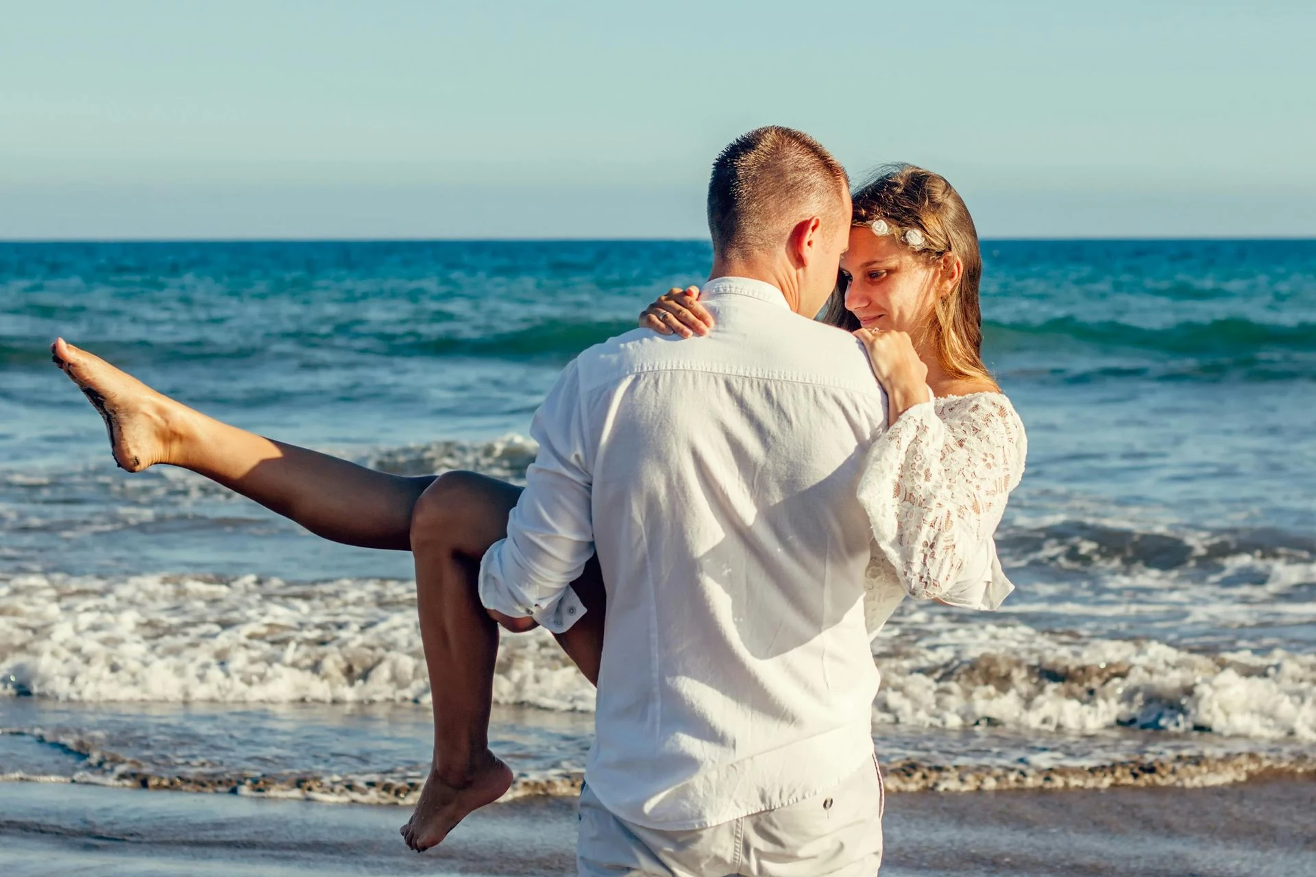 Man carrying woman at beach; turquoise water, white shirt, embrace, smile.