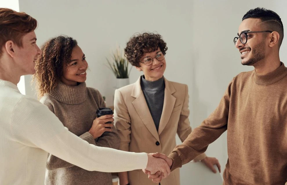 Four people in a room, two shaking hands, others smiling. Neutral tones, well-lit.