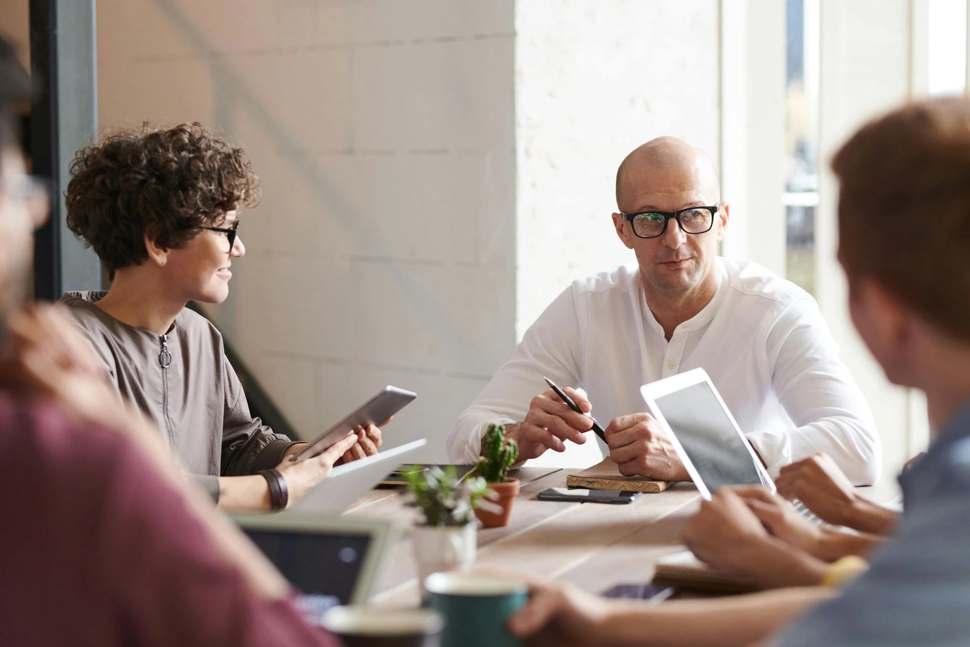 People in a meeting at a wooden table, using tablets and a pen. Bright office setting.