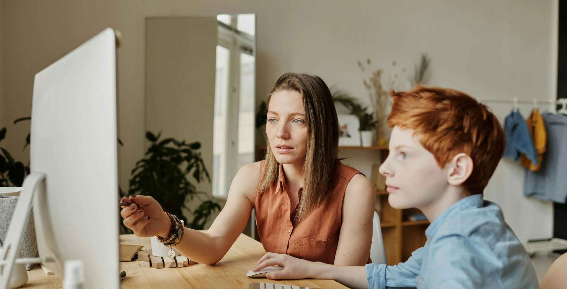Woman points at computer screen, assisting a boy with red hair at a desk.