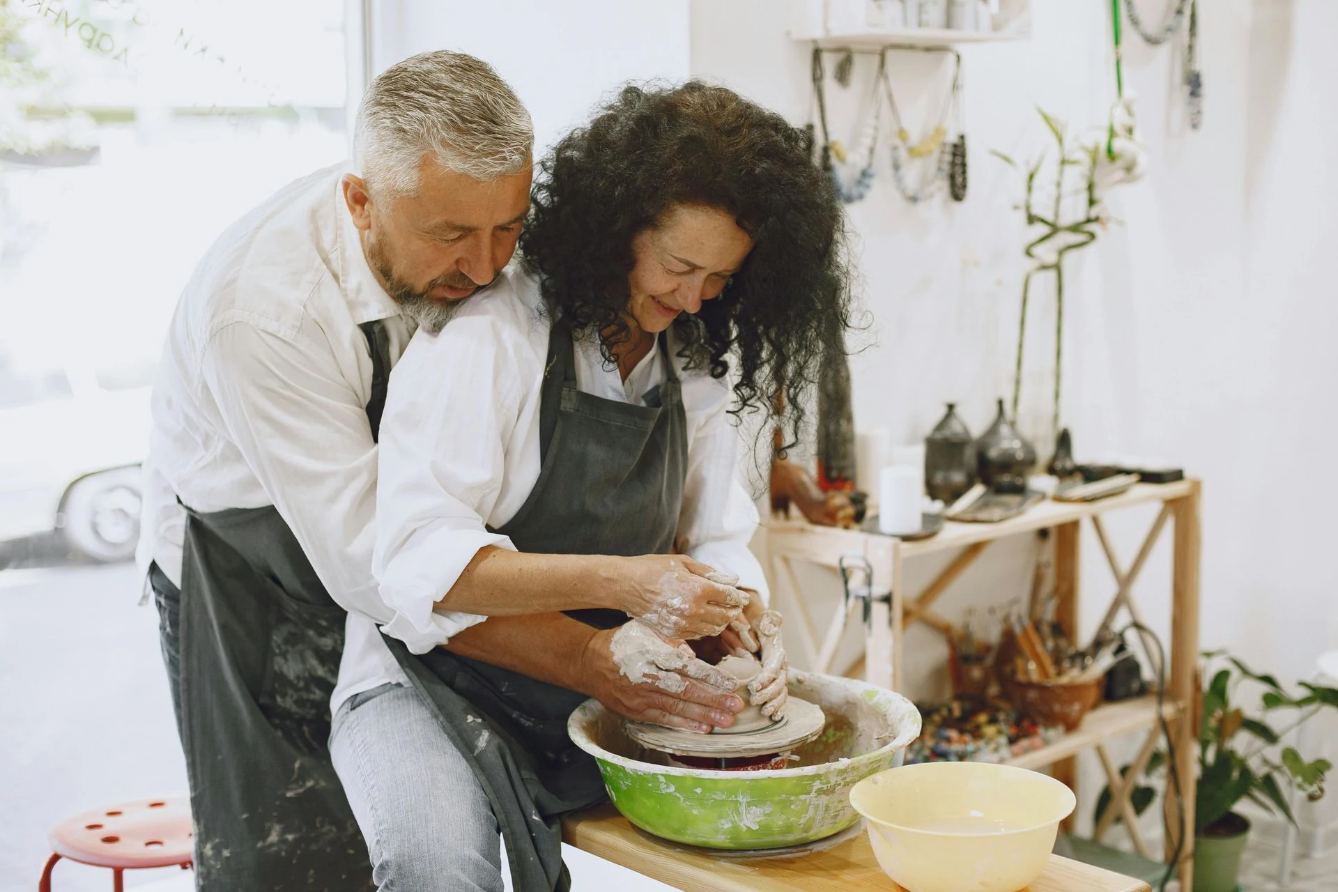 Man assisting woman with pottery wheel; they're working with clay, in an art studio setting.