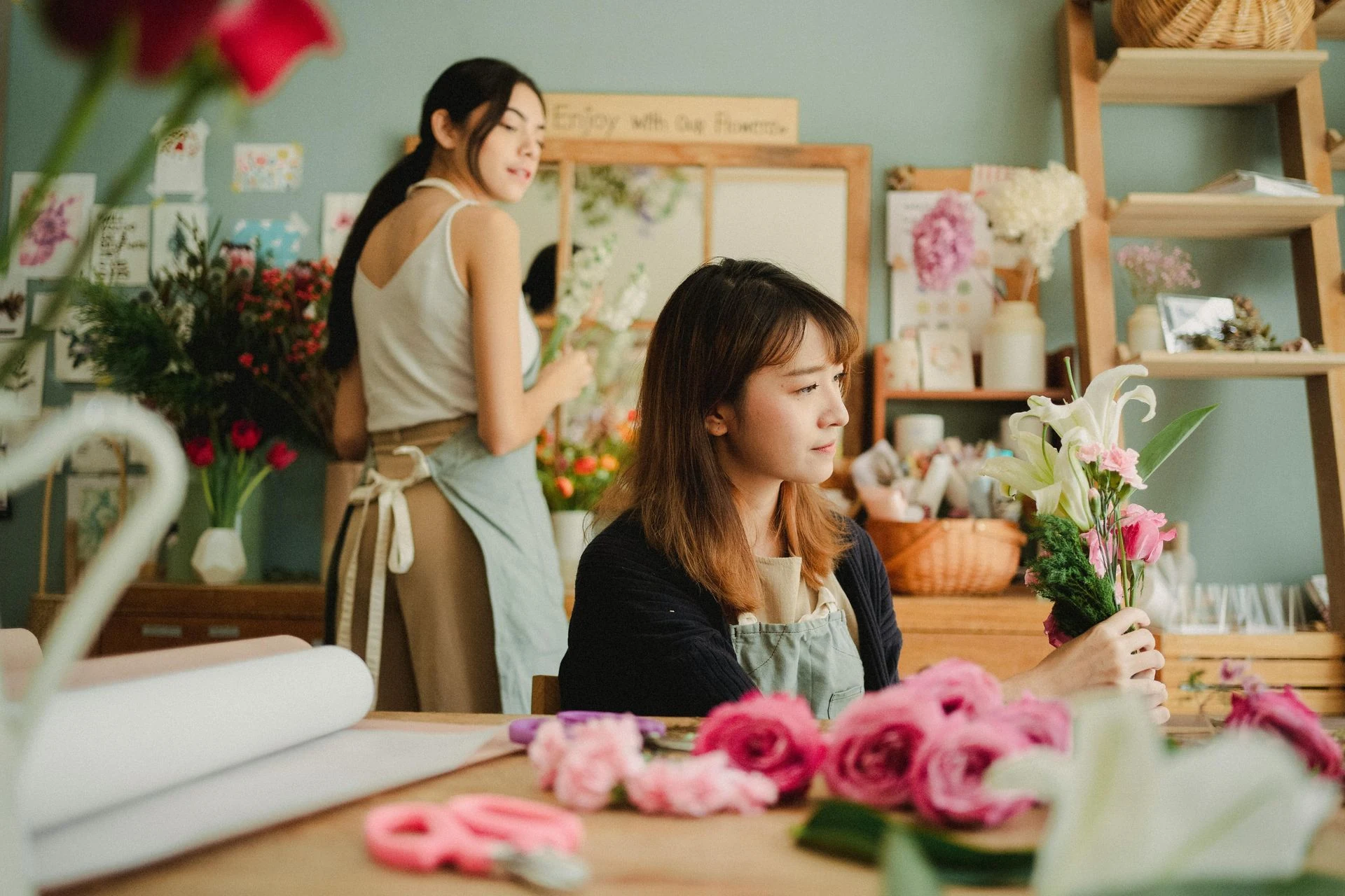 Two florists arranging flowers in a shop, one looking pensive, surrounded by blooms.