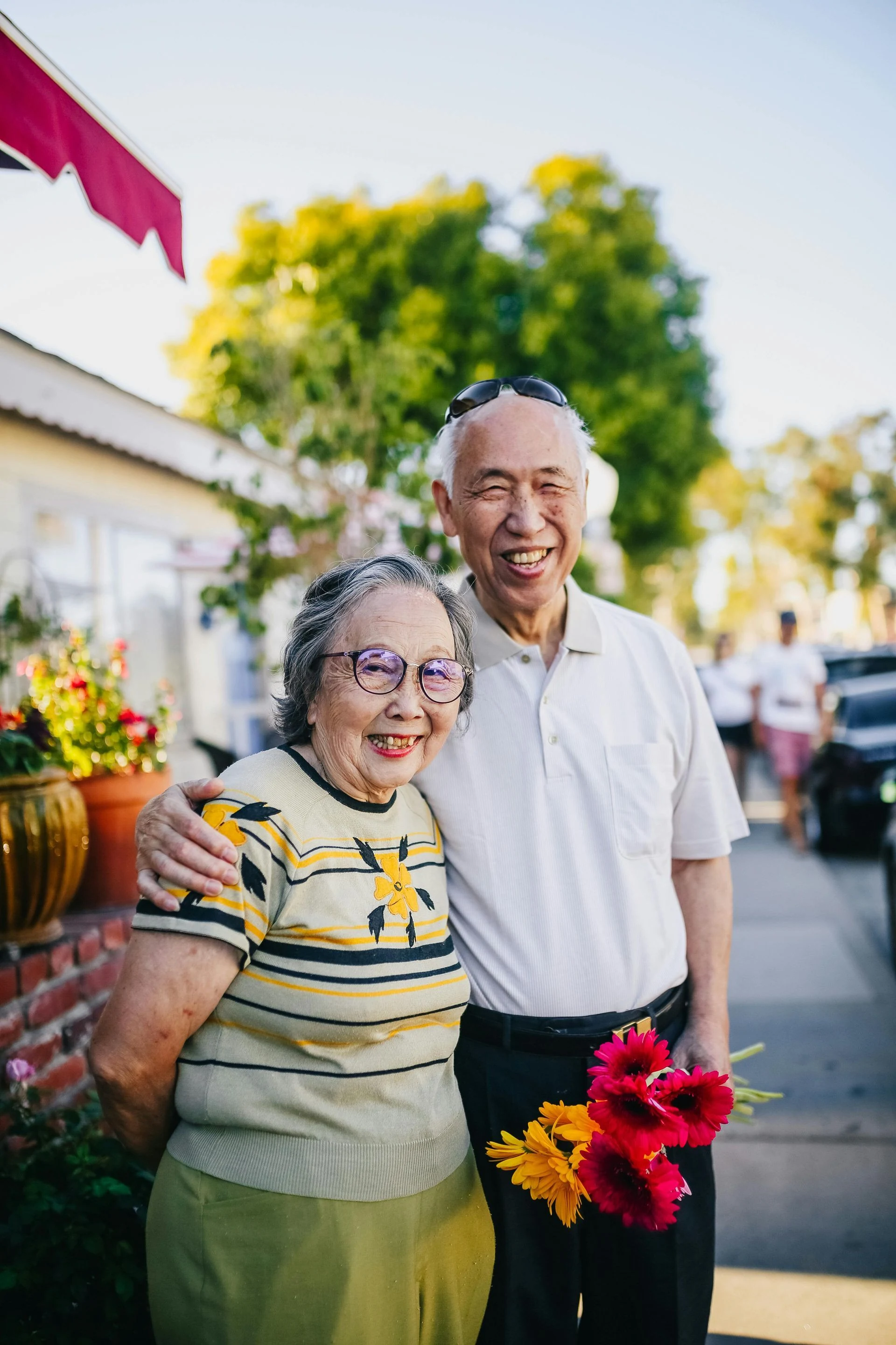 Elderly couple smiles for the camera, embraced, holding flowers outside a home on a sunny day.