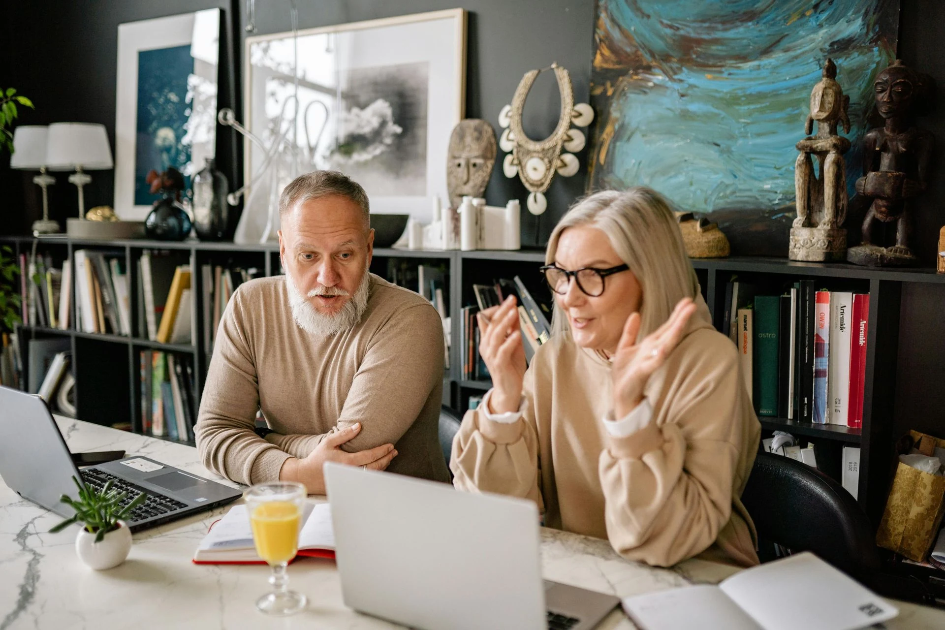 Man and woman at desk, working on laptops. The woman gestures, talking on a phone. Bookshelves and art in background.