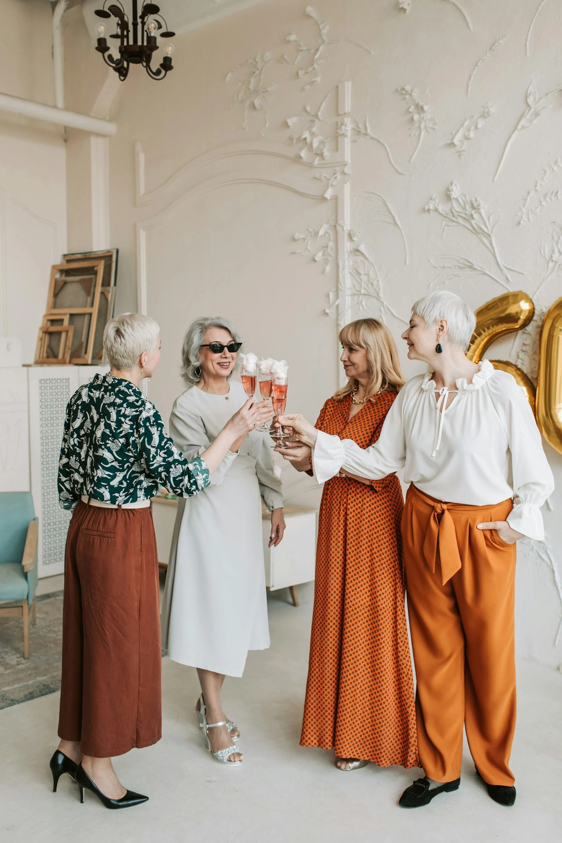 Four women in stylish outfits toast with champagne in a decorated room.