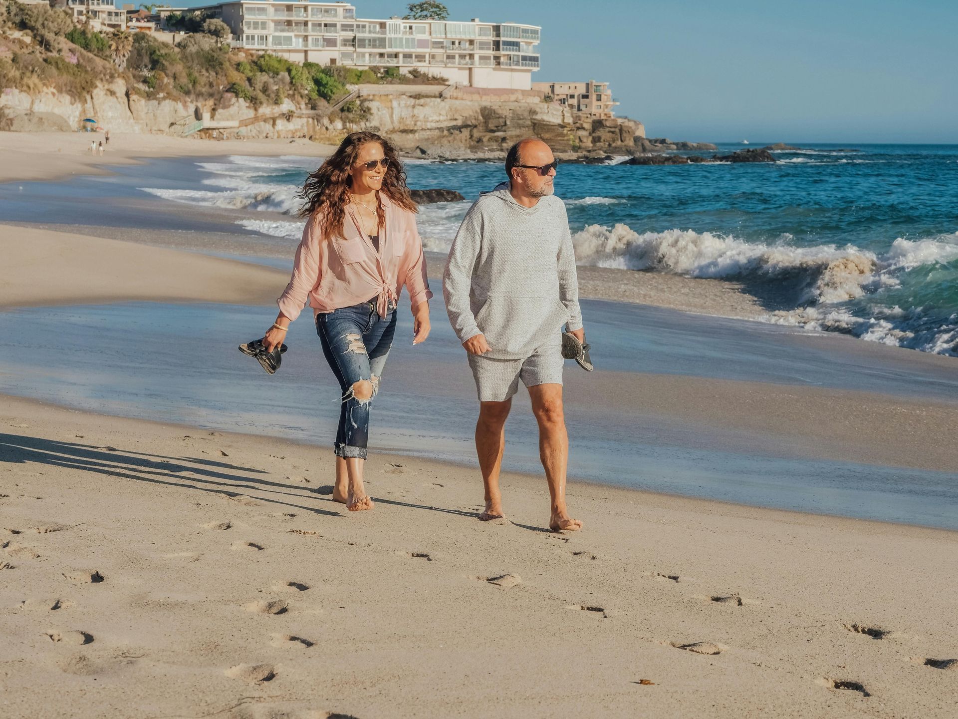 Couple walks barefoot along a beach. Woman in ripped jeans, man in a sweater; ocean waves and cliffside in background.