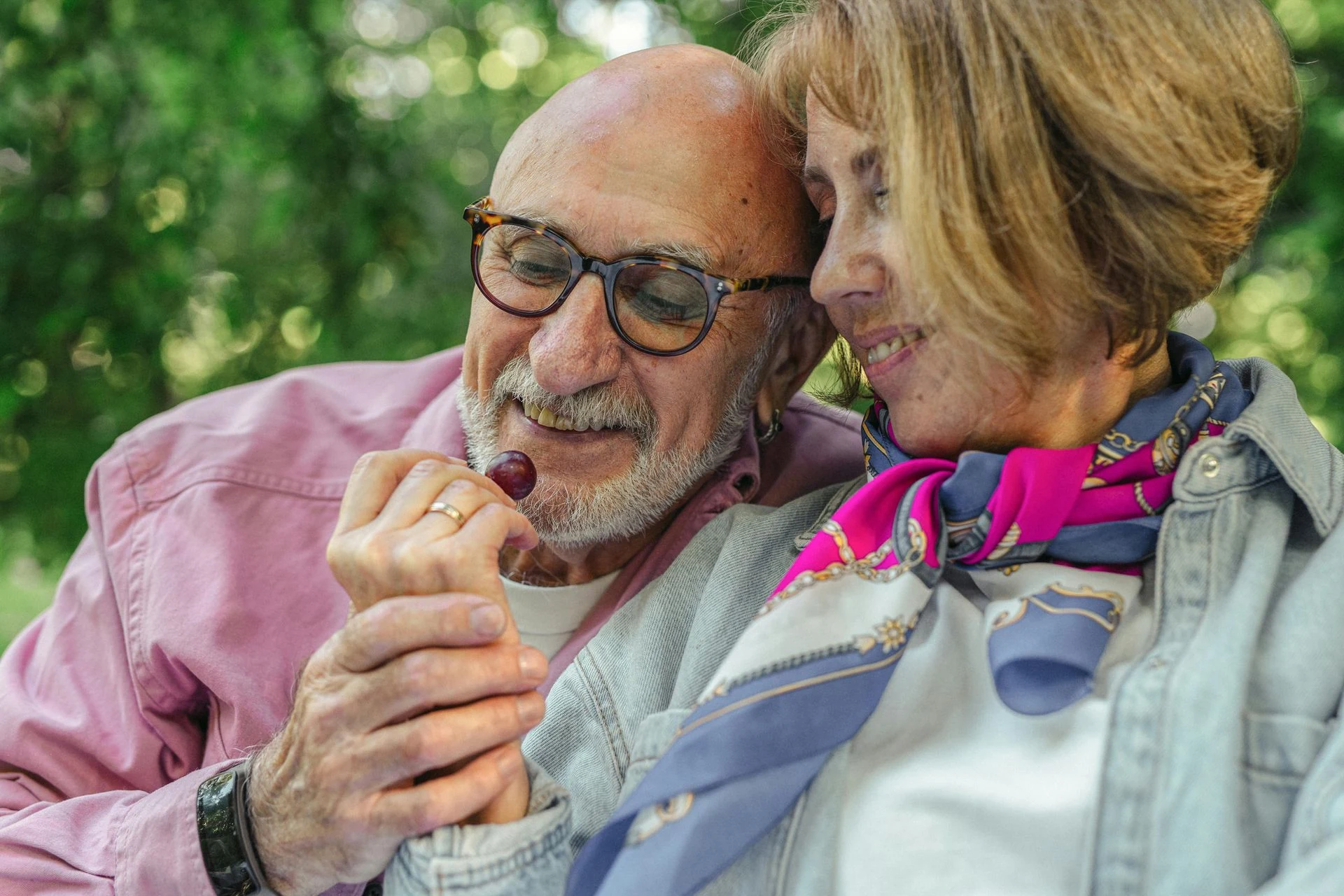 Older man and woman smile, sharing a cherry. They sit outdoors; she wears a scarf.