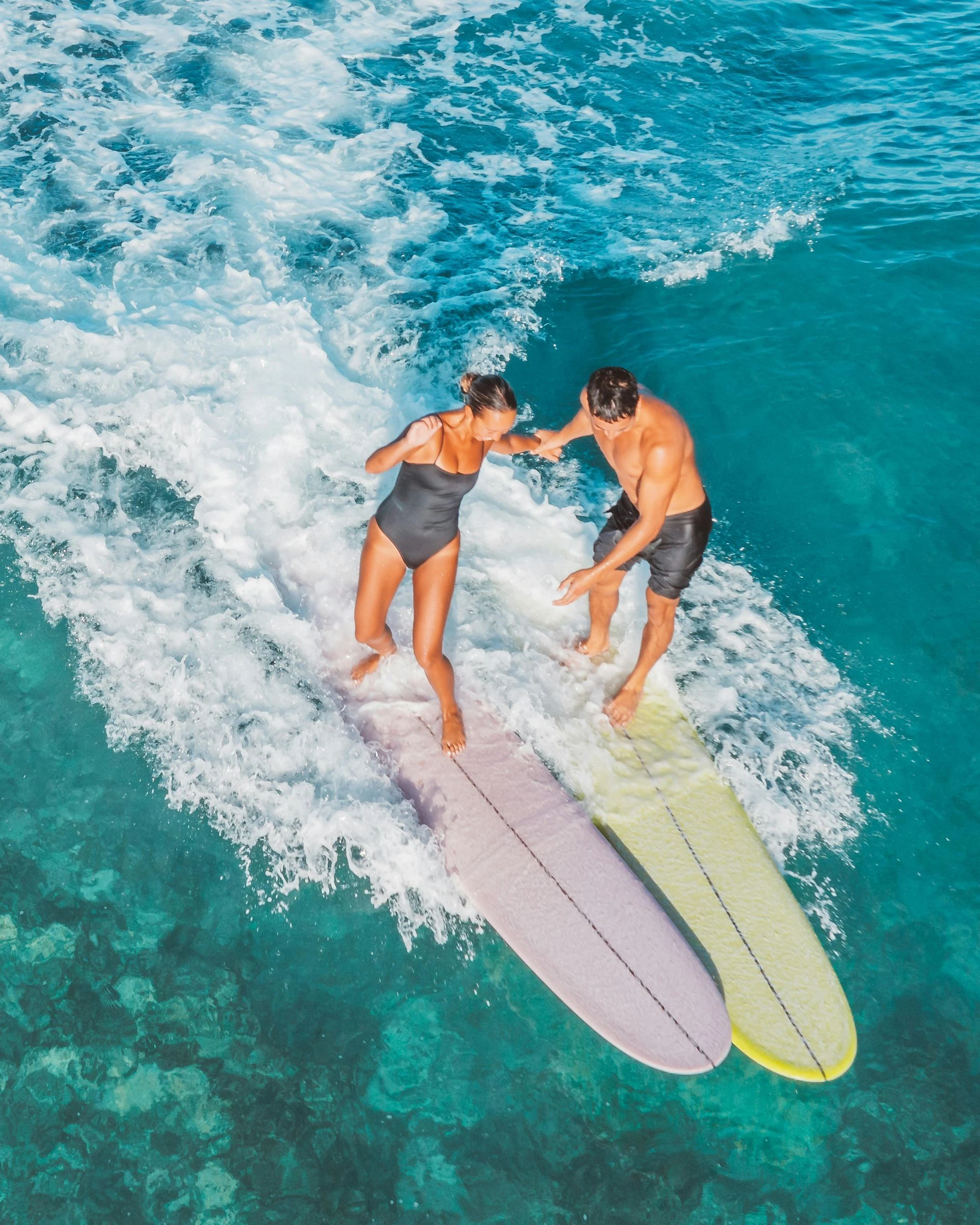 Two people surfing together on a blue ocean. The woman wears a swimsuit, and the man wears shorts.